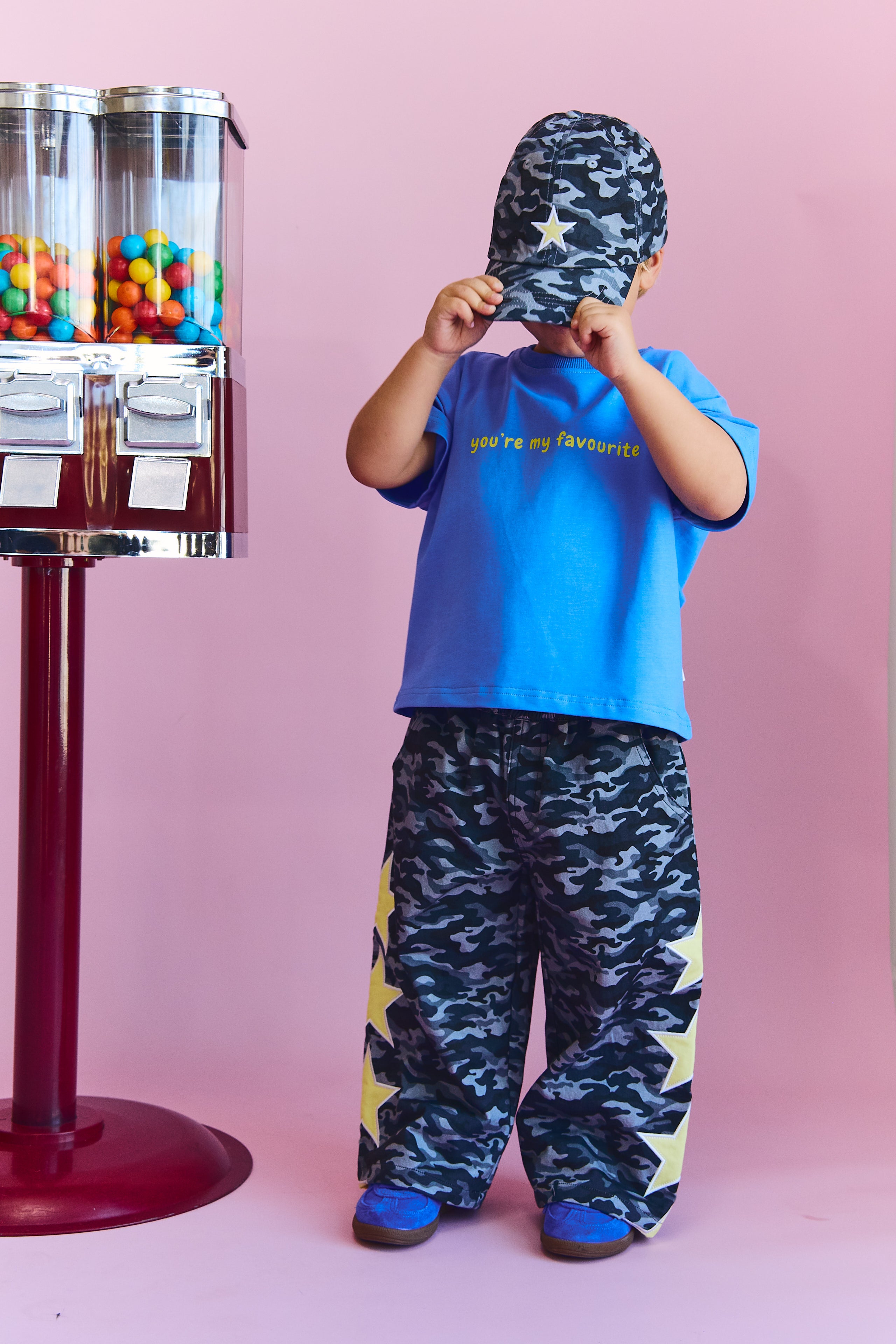 A toddler stands against a pink studio background wearing a blue t-shirt with yellow text, matching blue and black camouflage pants with yellow star accents, and a camouflage cap. A vintage-style ctwister machine with colorful gummy candies is visible on the left.