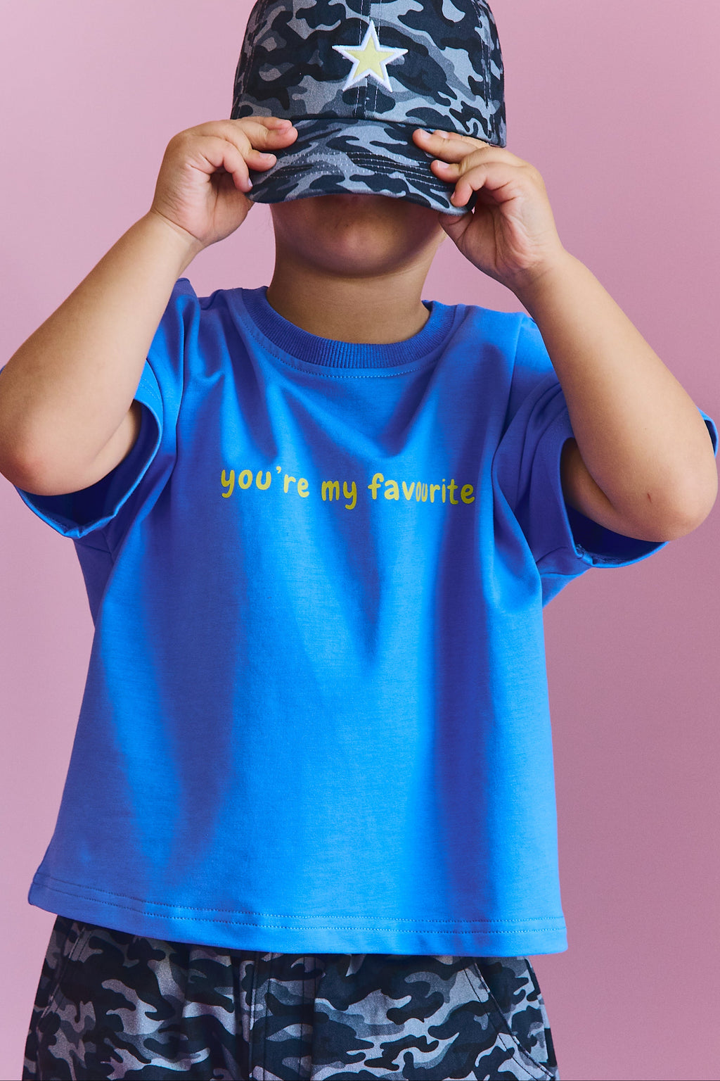 A child wearing a bright blue t-shirt with yellow text that reads 'you're my favorite' and matching dark blue camouflage shorts, holding a dark blue camouflage baseball cap in front of their face against a solid pink background.