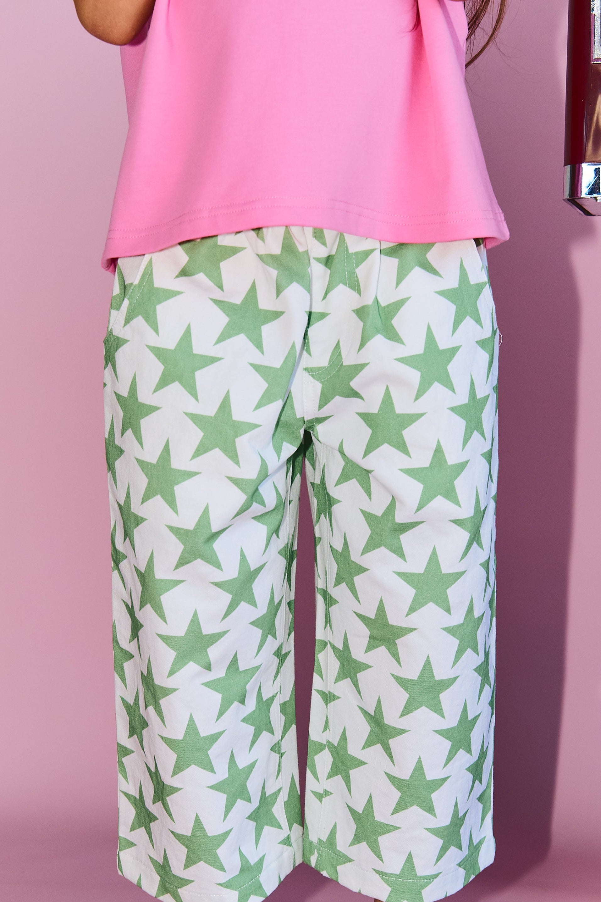A child wearing white cropped pants with a green star pattern, a pink top, and white sneakers with colorful laces, standing against a pink studio background.