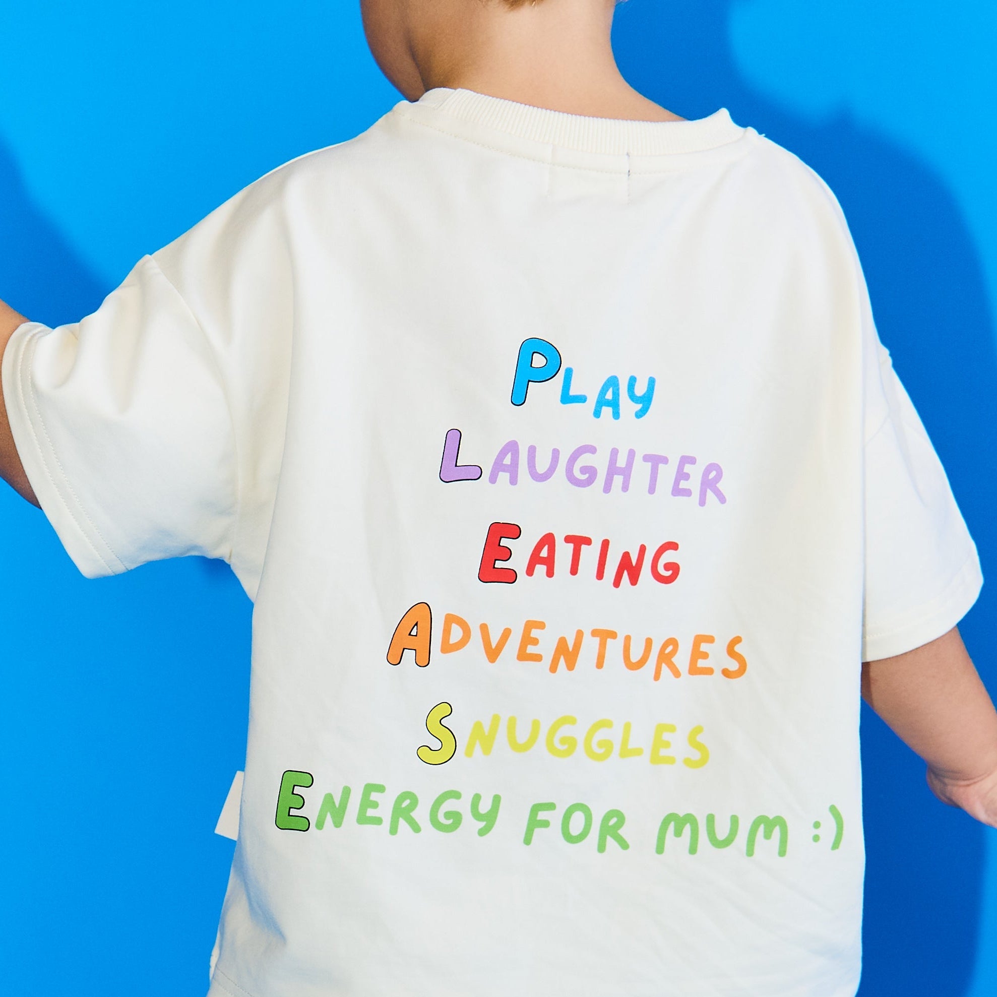 A child wearing a white t-shirt with colorful hand-written style text on the back and white pants with a green star pattern, posed against a bright blue background.