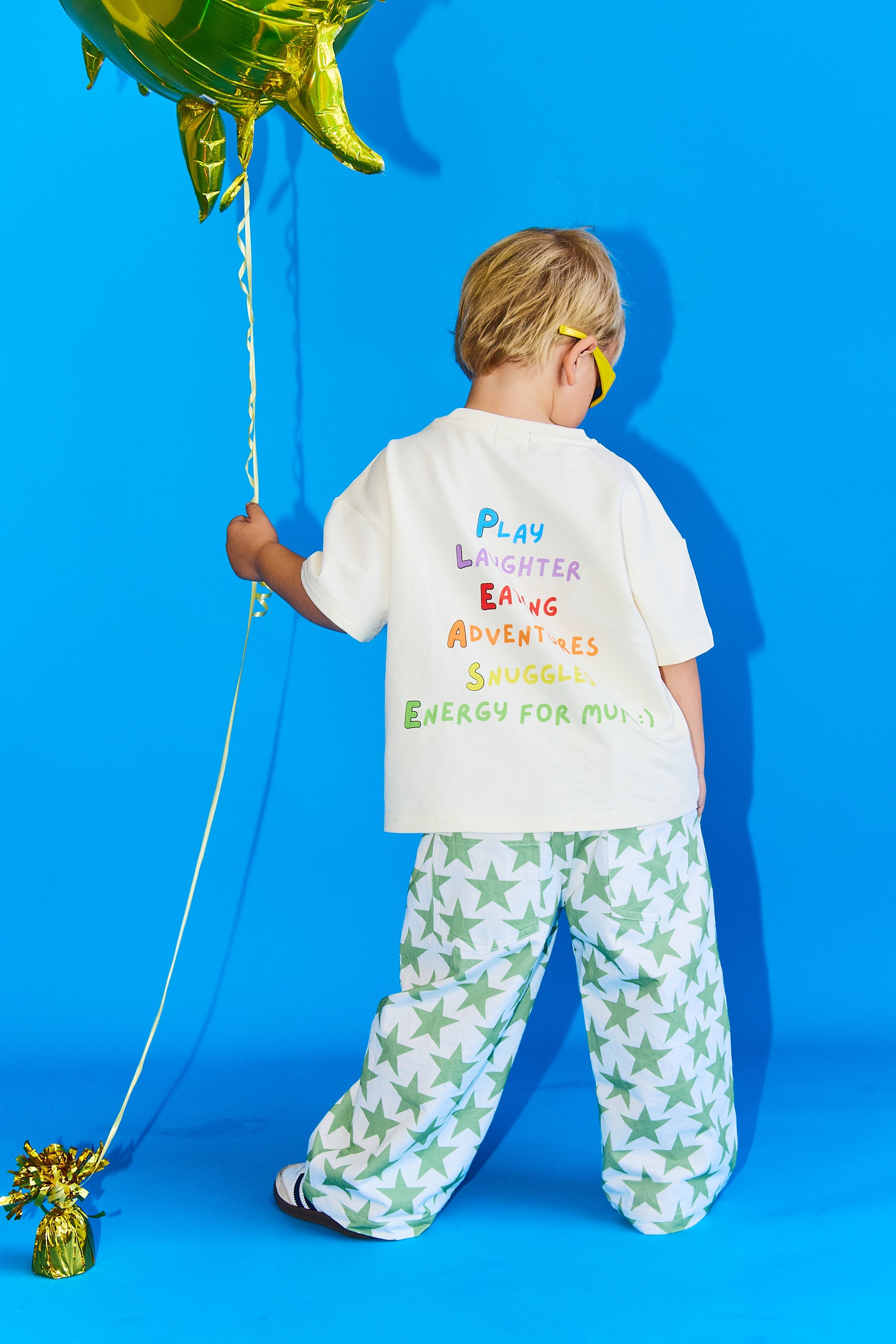 A child stands with their back to the camera wearing a white t-shirt with colorful text and green-and-white star-patterned pants, holding a gold foil balloon against a bright blue studio background.