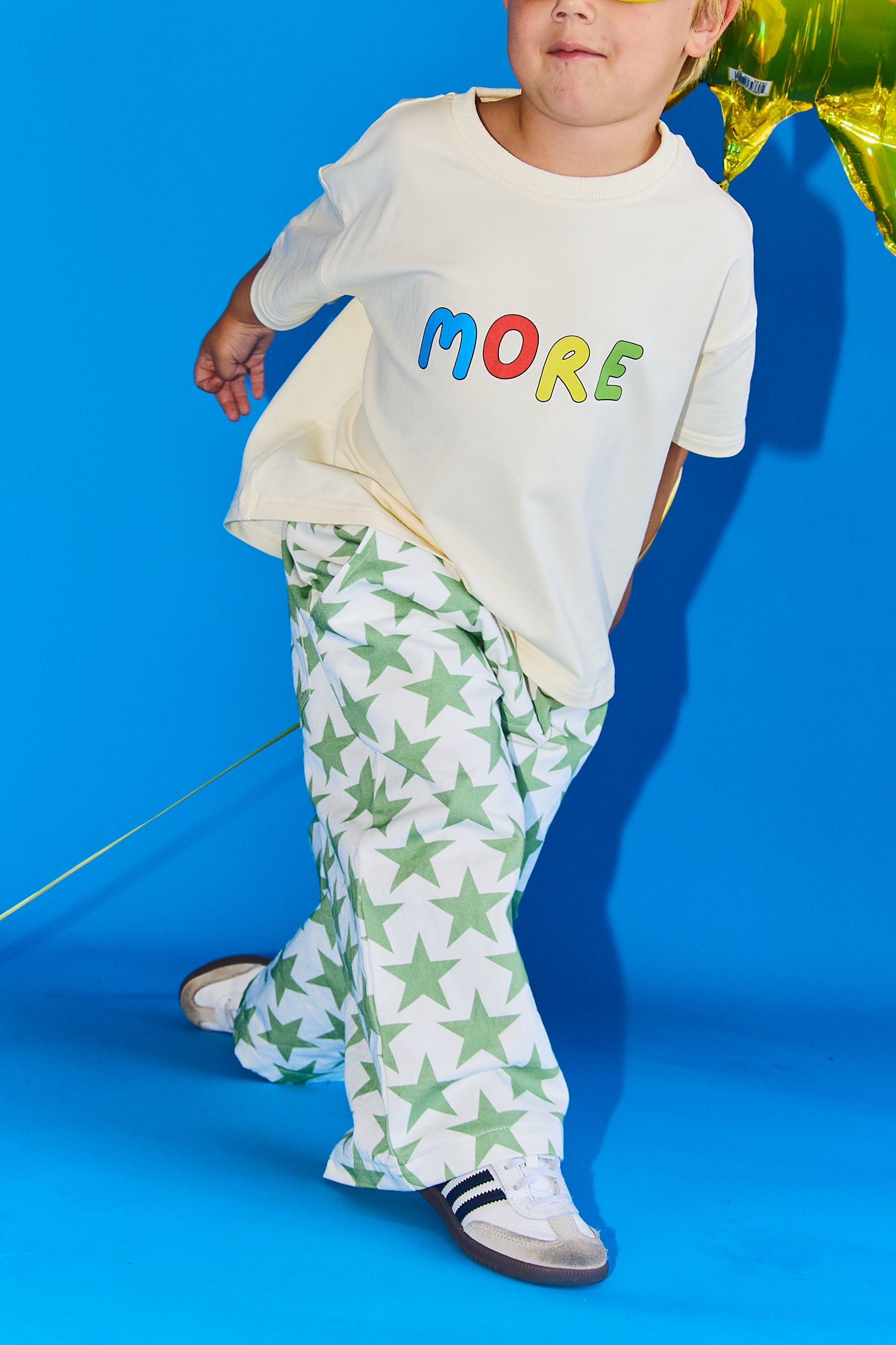 A young child wearing a cream t-shirt with the word 'MORE' in colorful letters and green-and-white star-patterned pants, posing against a bright blue studio background with a gold foil balloon.