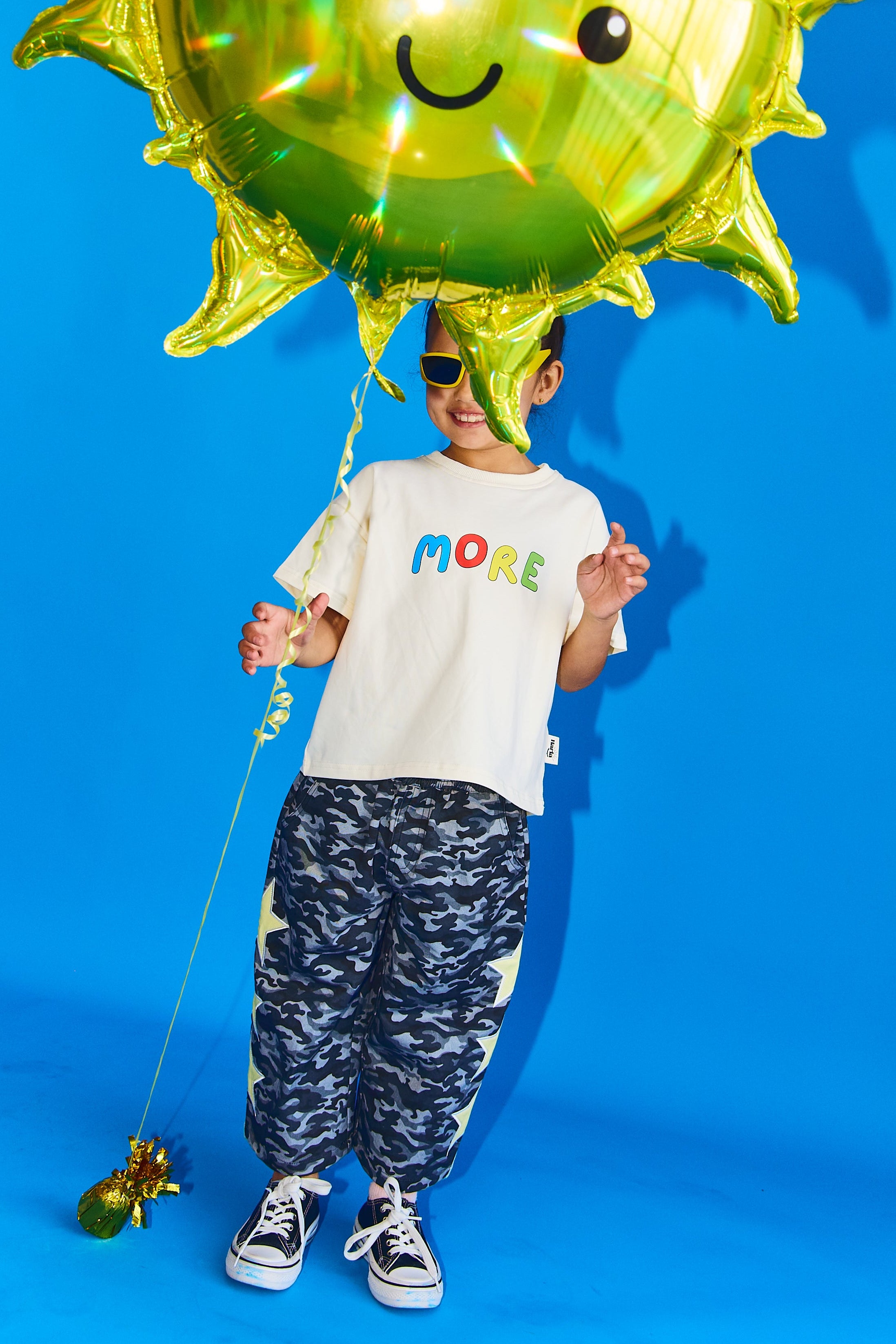 A child wearing a white graphic t-shirt with 'MORE' in colorful letters, blue and white camouflage pants, and black-and-white sneakers, holding a large metallic green sun-shaped foil balloon against a bright blue studio background.