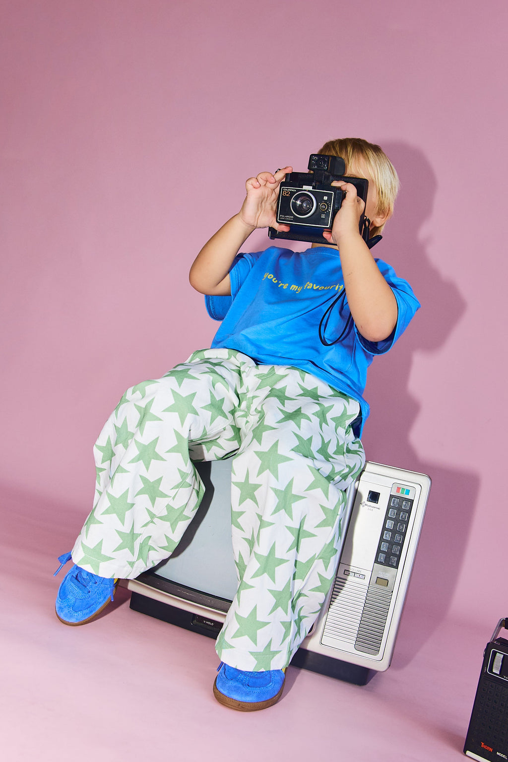 A child wearing a bright blue t-shirt and green-and-white star-patterned pants sits on a vintage television, holding a black camera up to their face. They are posed in front of a solid pink studio backdrop with retro electronic devices on the floor.