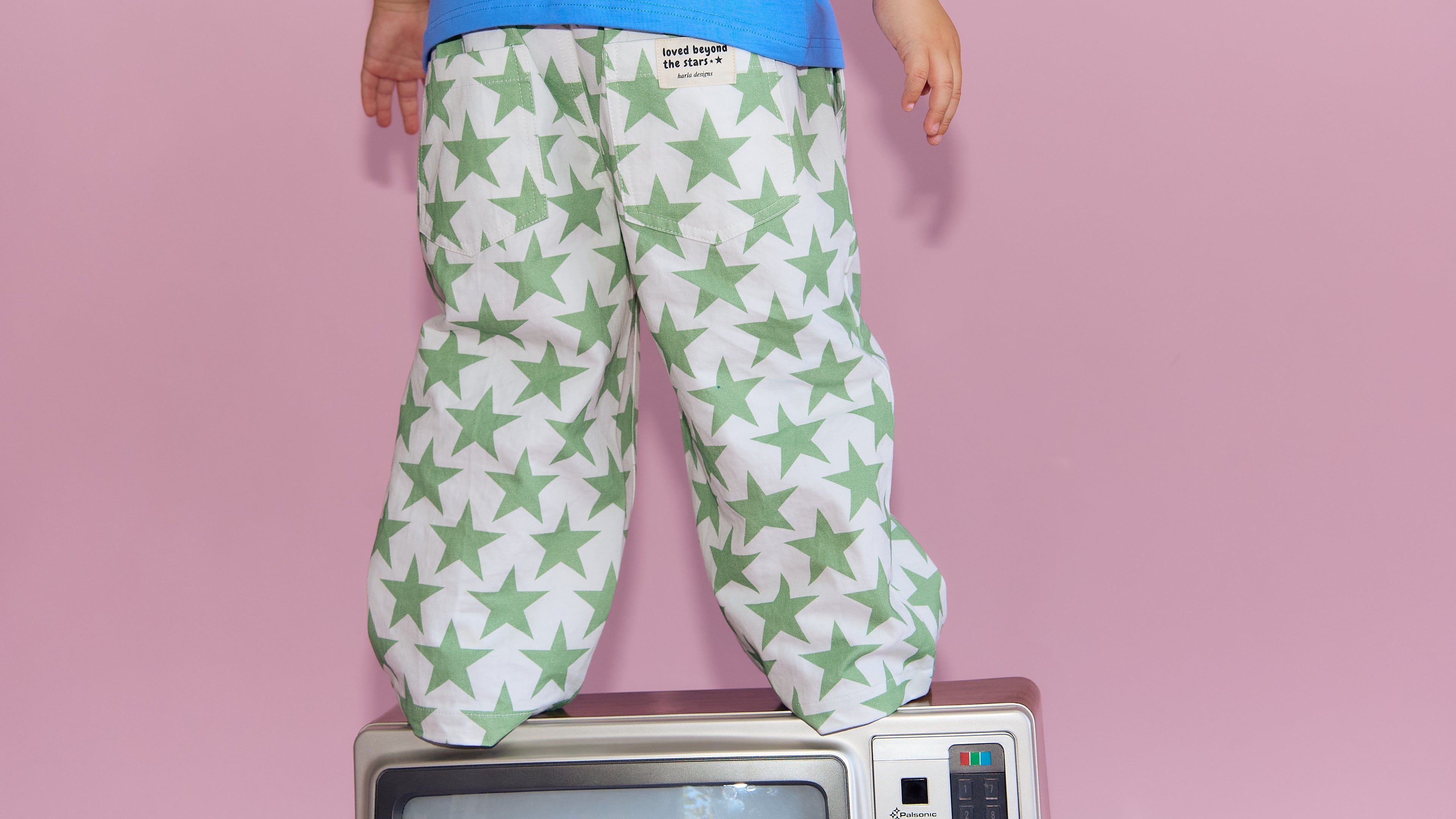 A child wearing white pants with a green star pattern and a blue shirt, standing on top of a vintage television against a solid pink background.