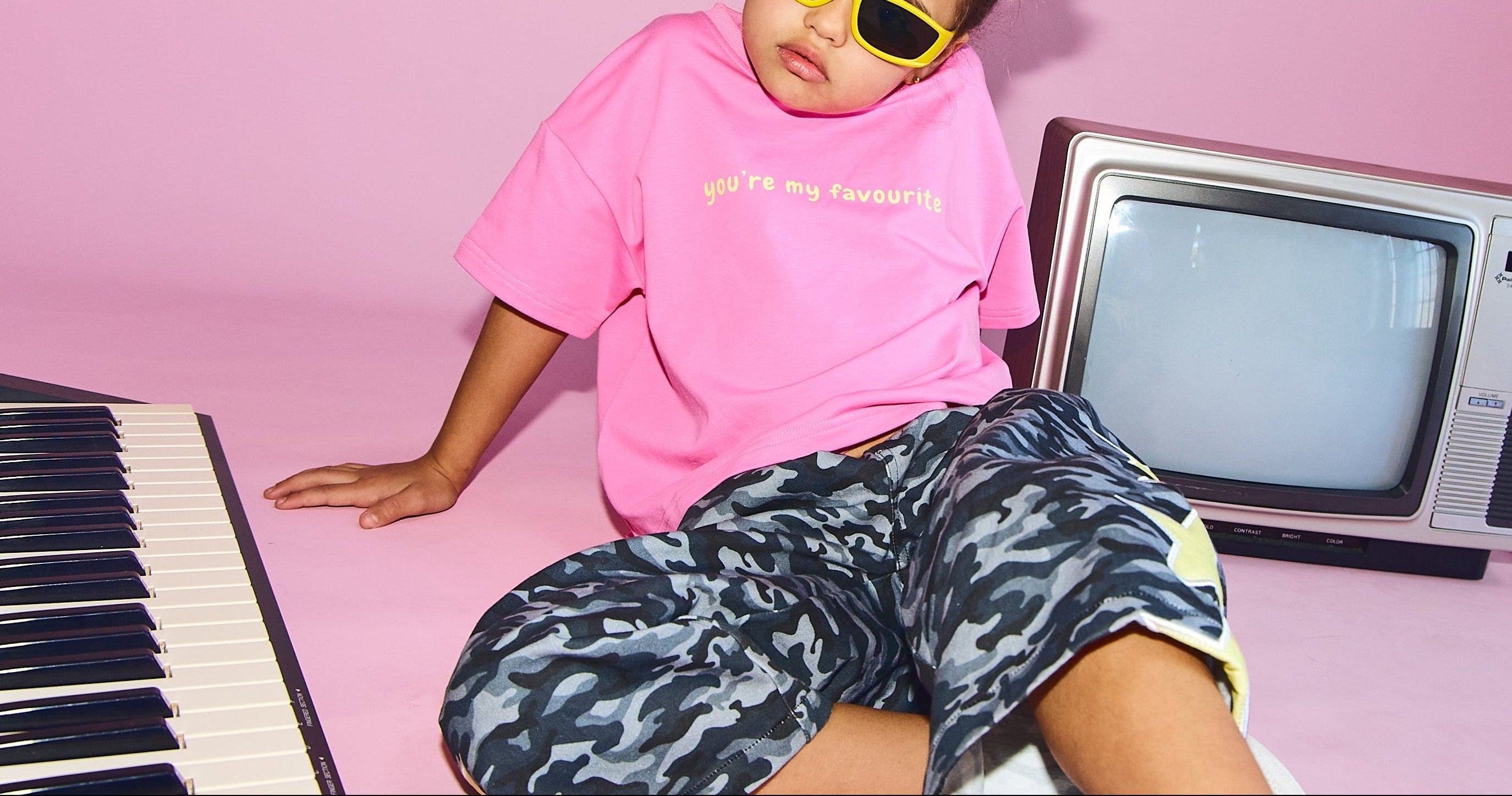A young girl with her hair in two buns sits on a pink floor wearing a pink t-shirt, gray and black camouflage shorts, and yellow sunglasses. She is posed with a vintage CRT television, a keyboard, and a tablet against a solid pink backdrop.
