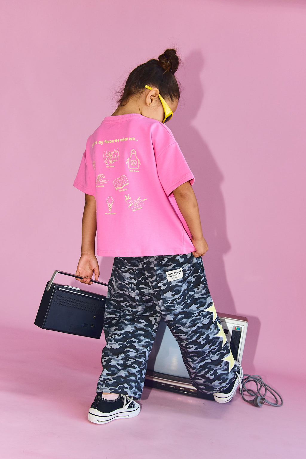 A child model from behind wearing a bright pink graphic t-shirt, black and gray camouflage cargo pants, and black sneakers, holding a vintage boombox against a solid pink studio backdrop.