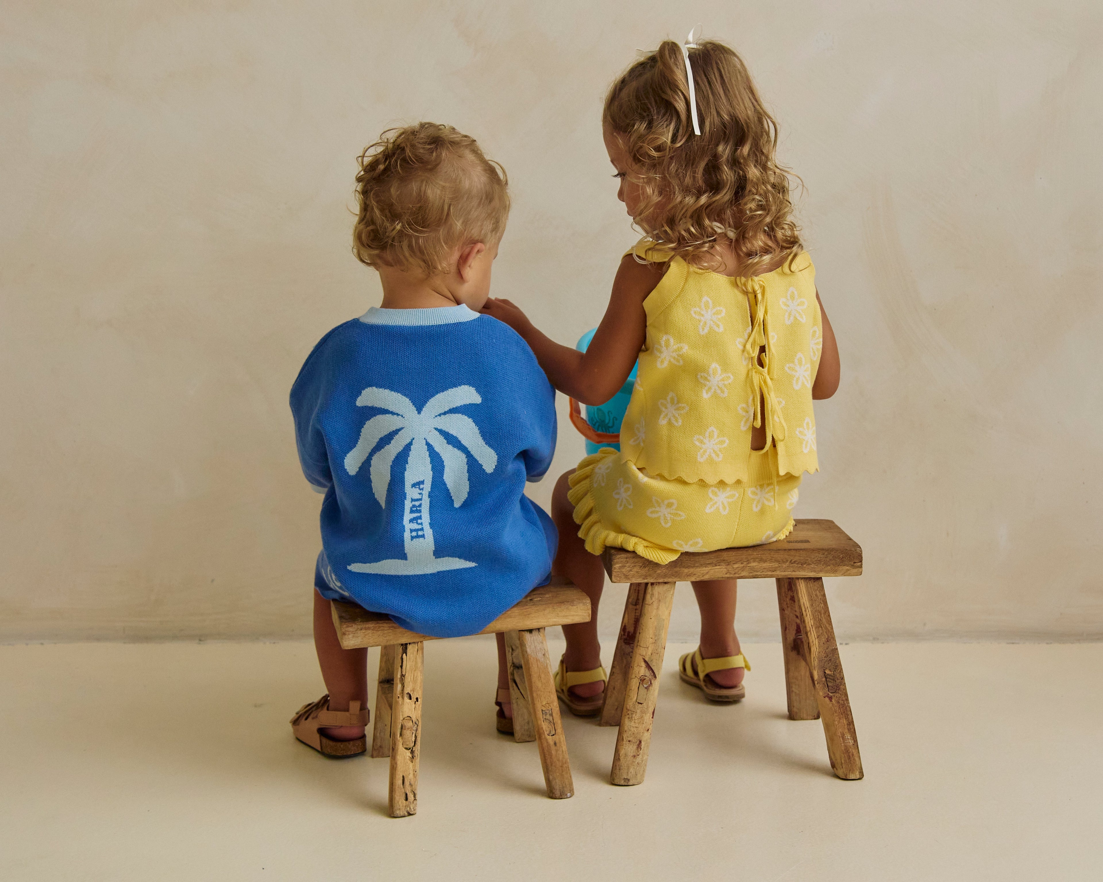 Two children sitting on wooden stools facing each other against a plain background